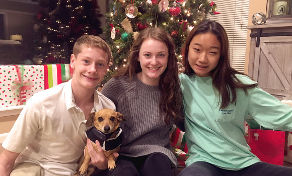 Three teenagers sitting together with a small dog in front of a decorated Christmas tree.