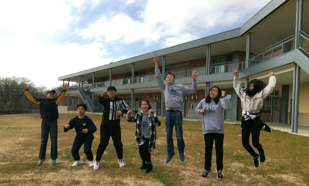 Students leaping in the air together on the lawn in front of a two-storey school building.