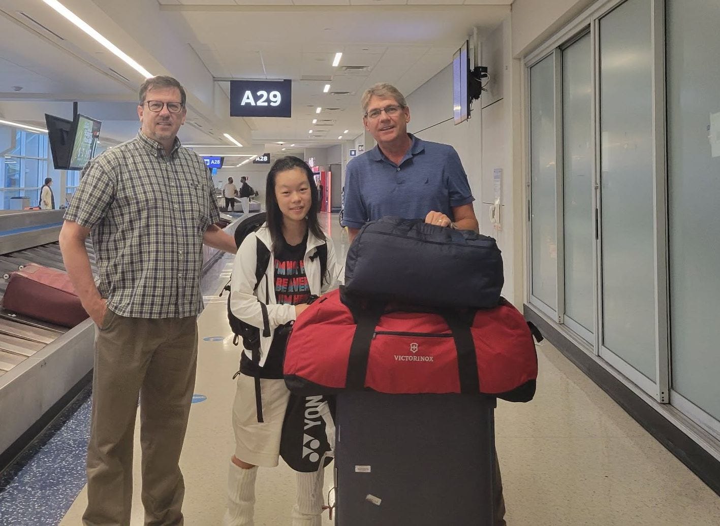 A student with luggage at an airport baggage carousel, greeted by two adults.